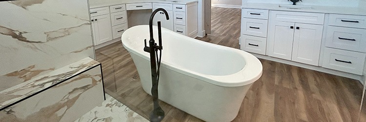 Spa-inspired custom bathroom featuring a freestanding soaking tub, bronze fixtures, marble stone walls, and white shaker cabinetry. Luxury interior design for Martin County homeowners by Masterpiece Builders.