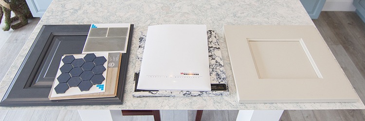 Cabinet door samples, tile swatches, and a Masterpiece Builders design catalog laid out on a marble countertop for renovation planning